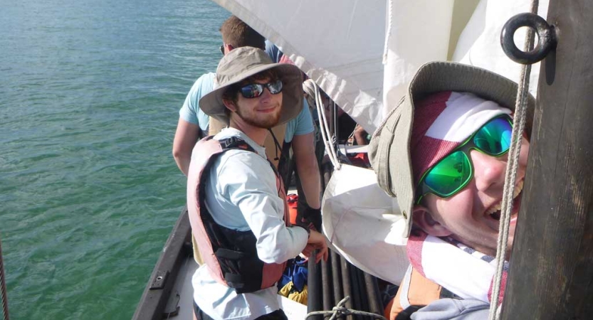 two people wearing hats, sunglasses and life jackets smile from a sailboat